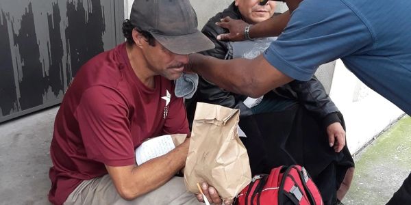 Three men sitting on a sidewalk with bags and a notebook.