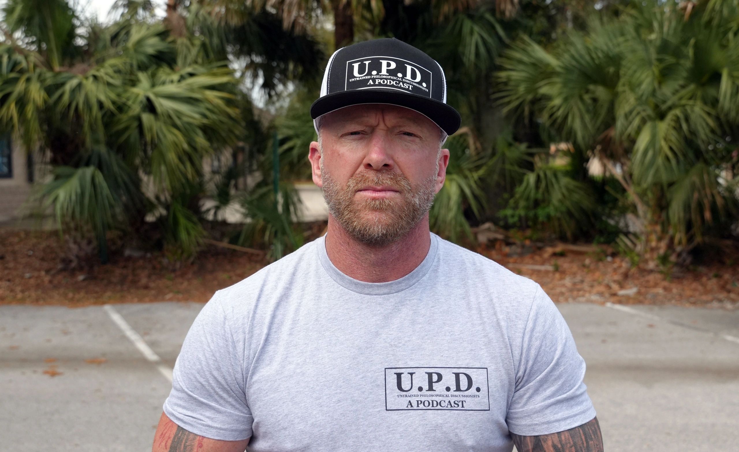 Man in U.P.D. podcast shirt and hat, standing outdoors with a serious expression.