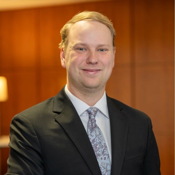 Smiling man in a suit and patterned tie against a warm wooden background.