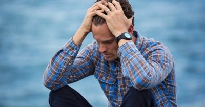 A man sits by the water, holding his head in distress.