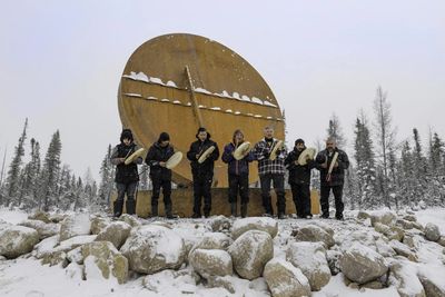 Seven people playing drums in snowy forest in front of a large drum structure.