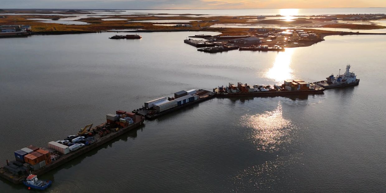 A long barge loaded with containers and vehicles floating on calm water at sunset.