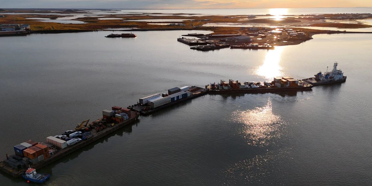 A long barge loaded with containers and vehicles floating on calm water at sunset.