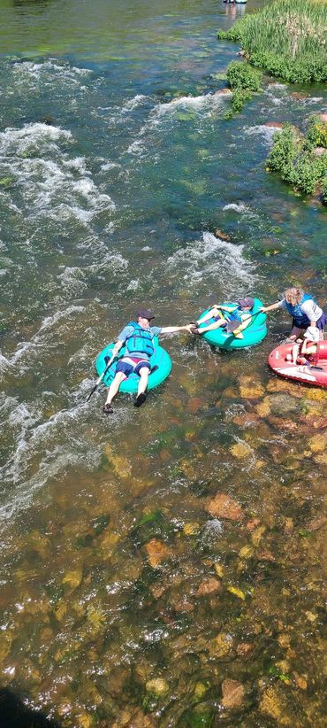 People tubing down a clear, rocky river, reaching out to each other.