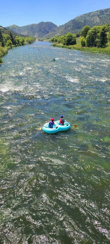 Two people rafting down a sunny river surrounded by mountains and trees.