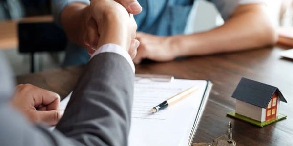 Two people shaking hands over a contract with a model house and keys on the table.