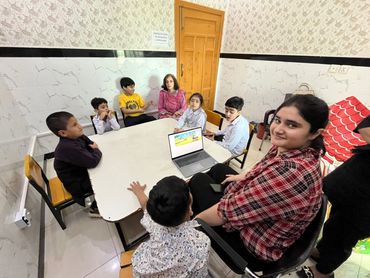 Group of children and two women gathered around a table with a laptop in a room.