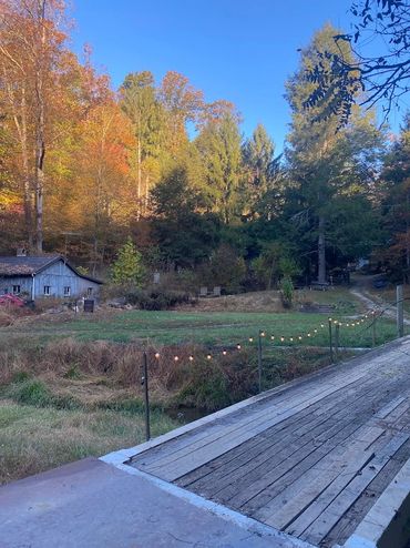 Rustic wooden bridge over a small creek with autumn foliage and a cabin in the background.