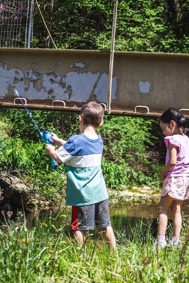 Two children fishing by a small creek under a metal bridge on a sunny day.