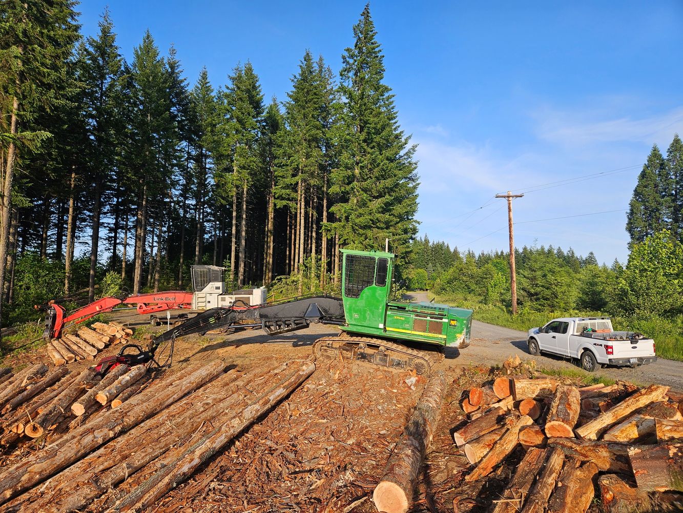 Logging machinery and a pickup truck near a forest with cut logs.