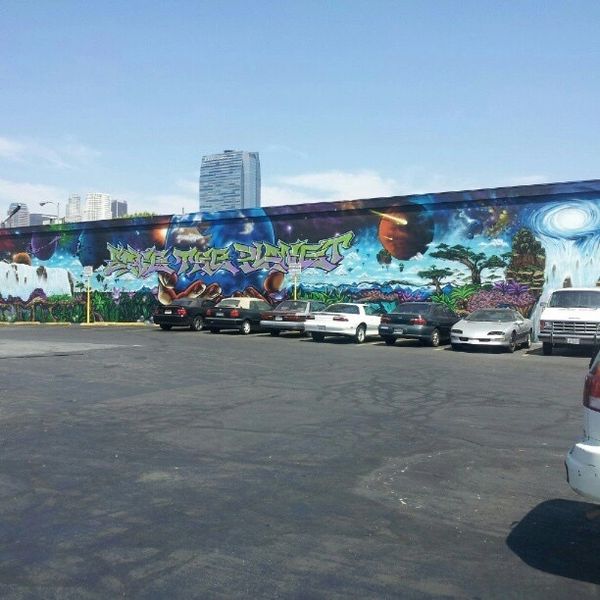 A colorful mural with space and nature themes on a long wall behind parked cars.