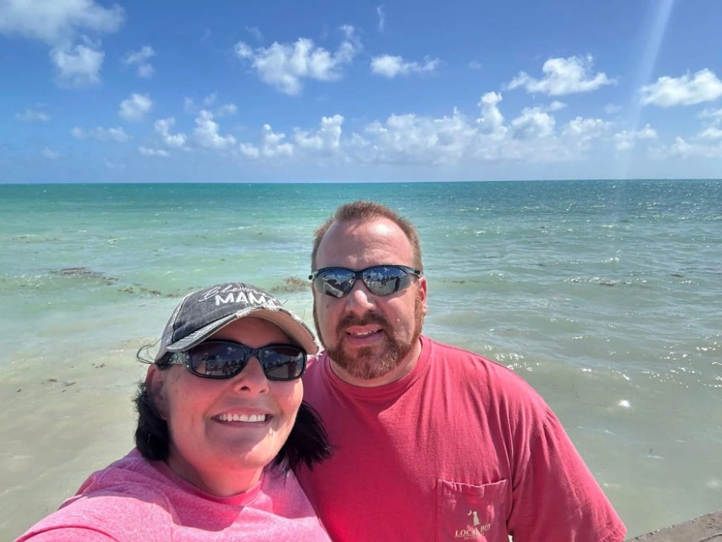 A happy couple wearing sunglasses takes a selfie at the beach under a bright blue sky.