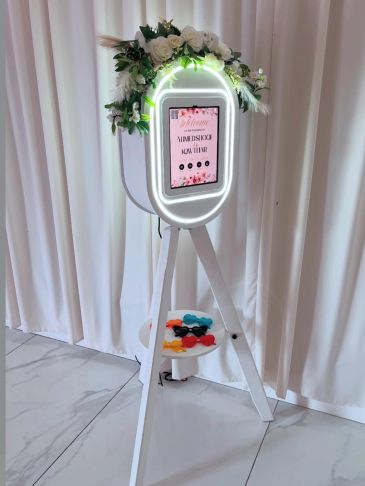 Elegant photo booth with floral decor and colorful props on a white tripod stand.