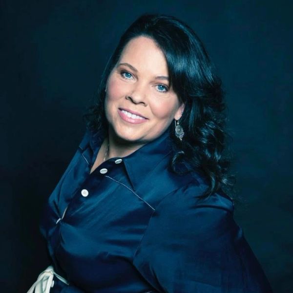 Woman with dark hair and blue shirt smiling against dark background.