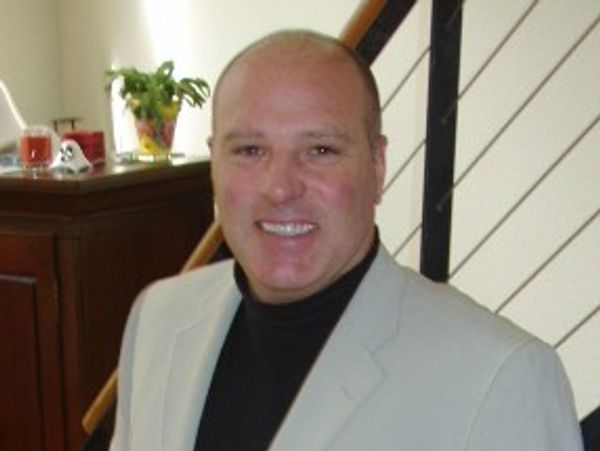 A man in a light-colored blazer smiling indoors.