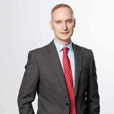 Man in a dark suit with a red patterned tie posing confidently.