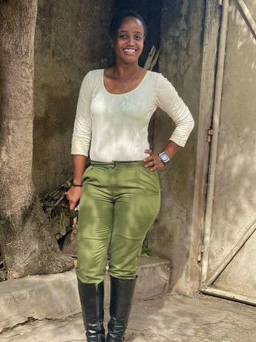 A woman in a white top and green pants standing outdoors near a tree and metal gate.
