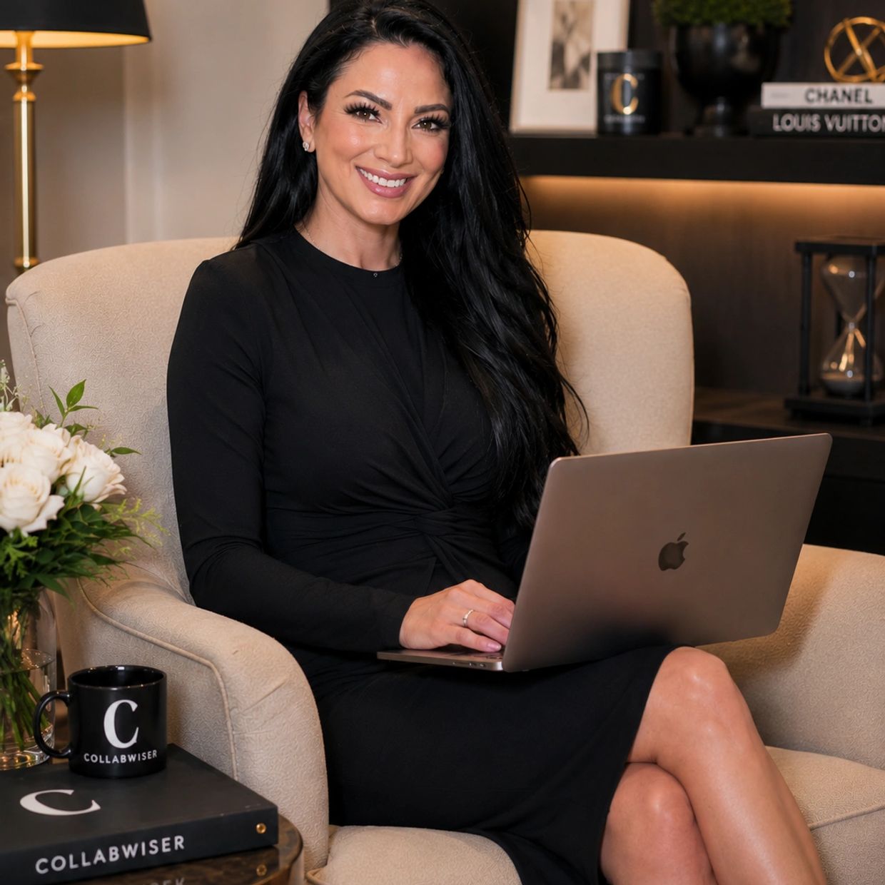 Woman in black dress sitting on a beige armchair with a laptop, smiling.