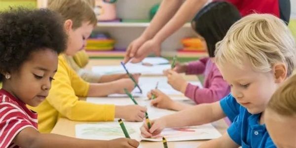 Children coloring pictures at a classroom table with a teacher's guidance.