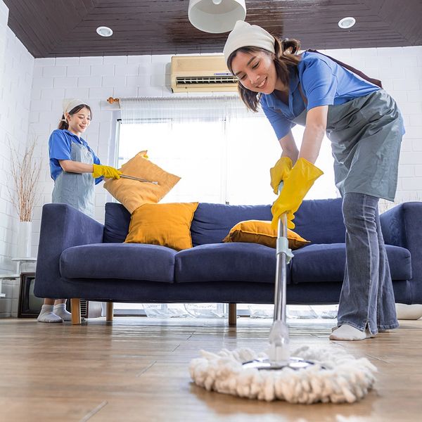 Two women cleaning a living room, mopping the floor and fluffing cushions.