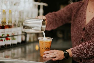 A person pours milk into iced coffee at a café counter.