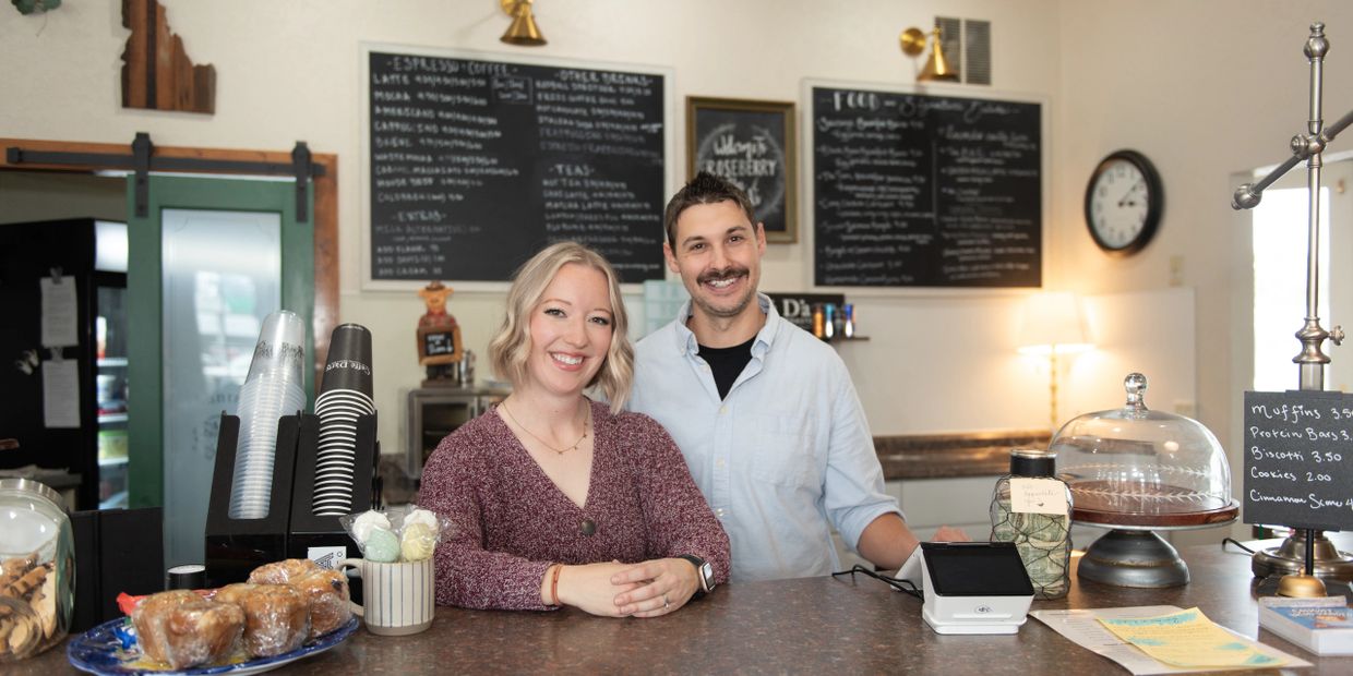 Two smiling baristas stand behind a coffee shop counter with pastries and menu boards.