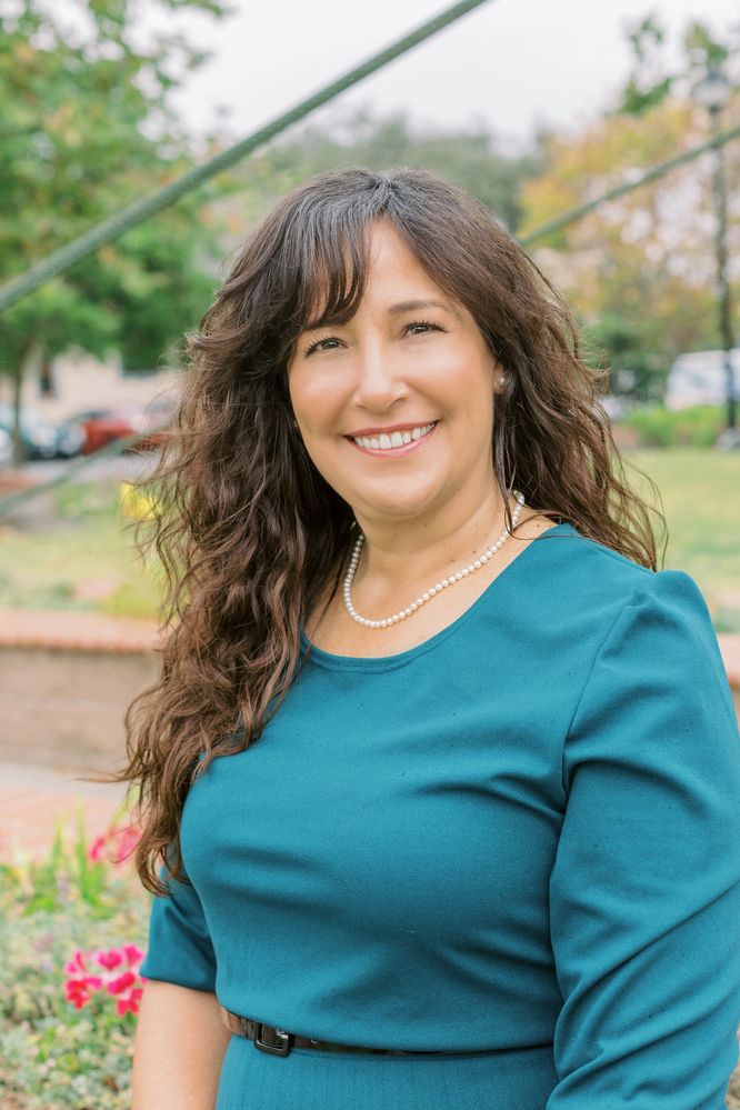 Smiling woman in teal dress with pearl necklace outdoors.