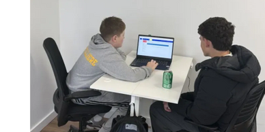 Two young men working on a laptop at a white desk with a soda can.