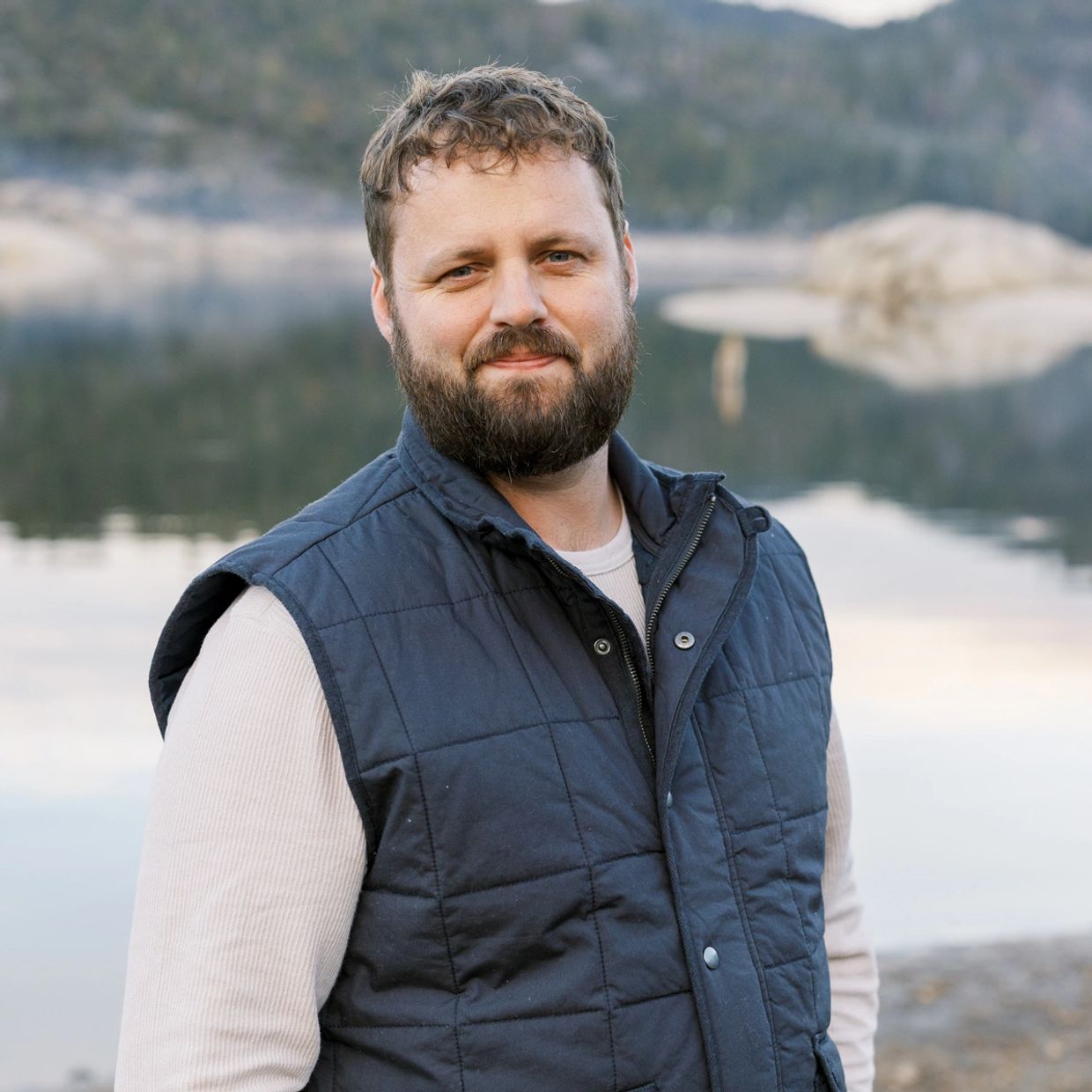 Man with beard in a navy vest standing by a calm lake.