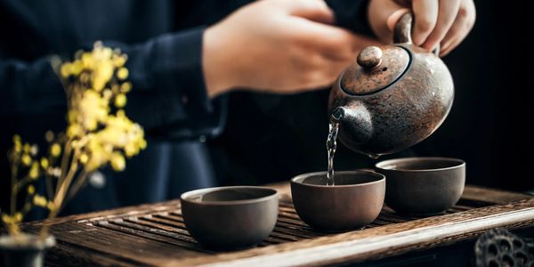 Person pouring tea from a rustic teapot into ceramic cups on a wooden tray.