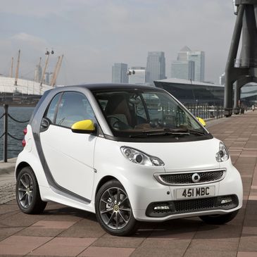 Compact white Smart car with yellow side mirrors parked near a waterfront.