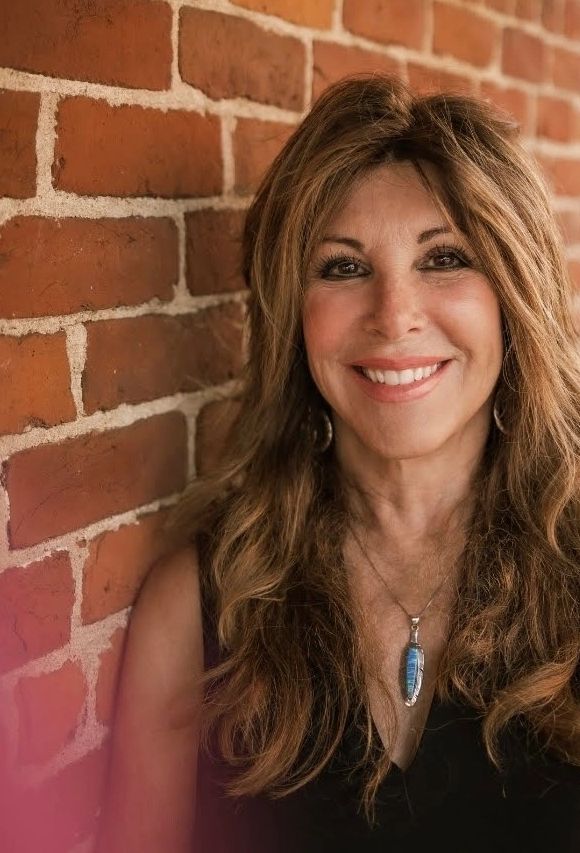 Smiling woman with long hair leaning against a brick wall.