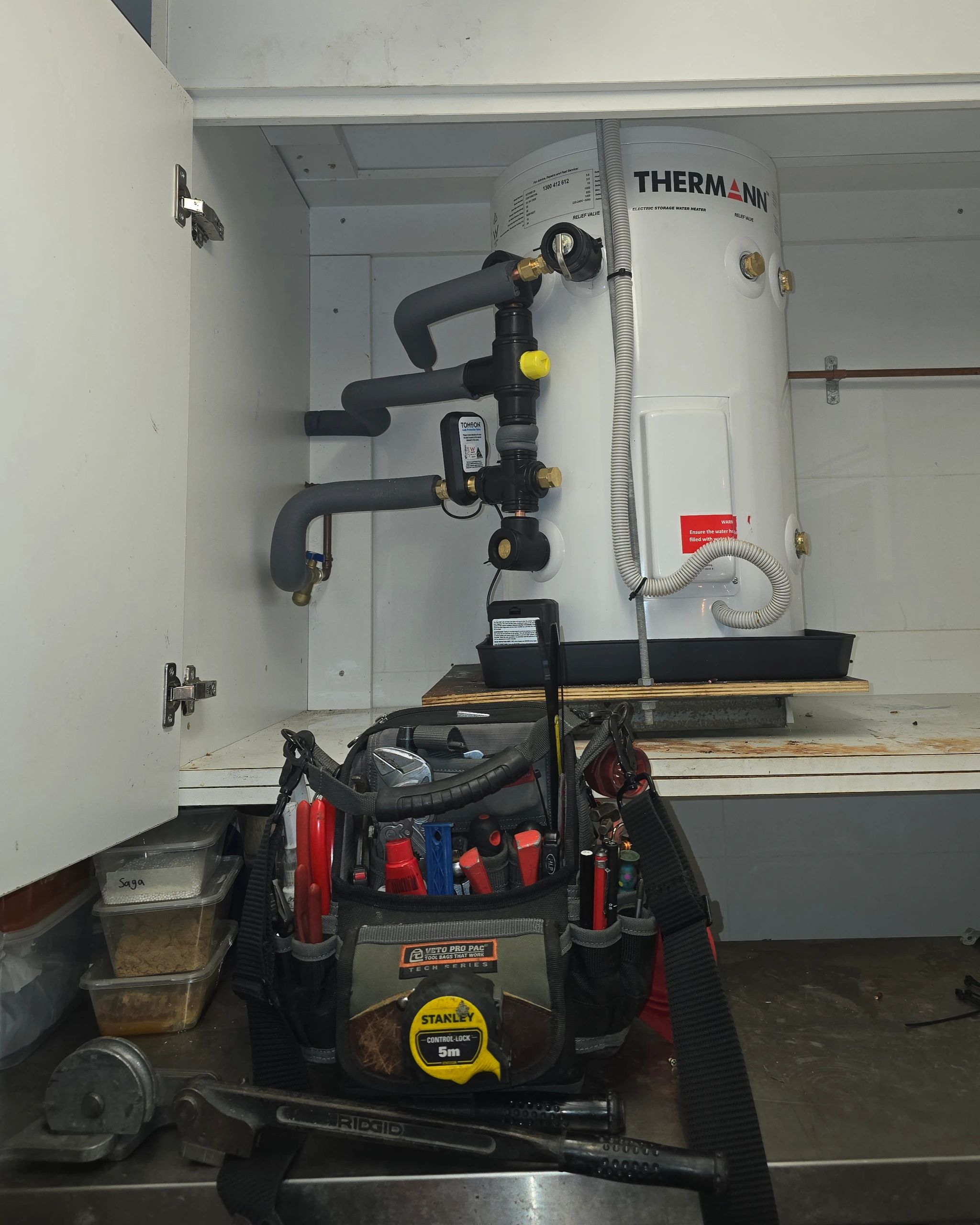 Tool bag and plumbing tools in front of a water heater inside a cabinet.