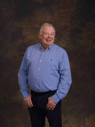 Smiling older man in a blue shirt posing against a dark, textured background.