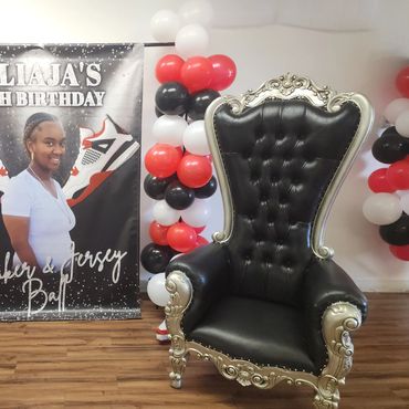 Black tufted silver throne displayed at a 16th birthday celebration with basketball-themed balloon arch and custom backdrop for guest of honor seating.