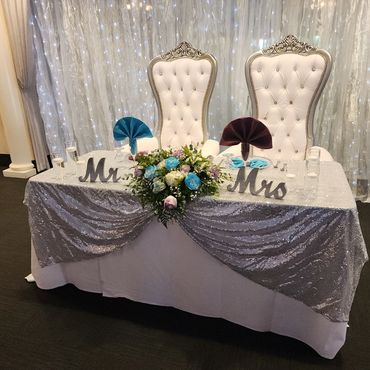 Pair of white and silver throne chairs set behind a sequin sweetheart table with Mr. & Mrs. signs and florals. (124 characters)