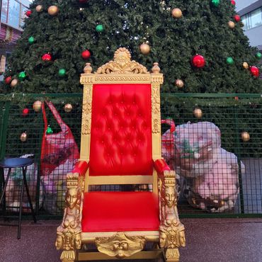 Red and gold throne placed in front of giant Christmas tree at outdoor event for public photo opportunities.