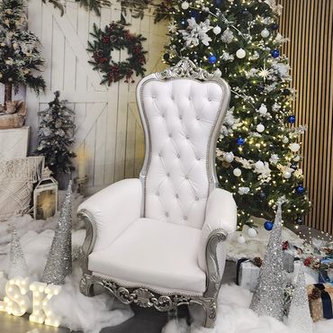 White and silver throne set beside a decorated Christmas tree with snow-like floor and light-up display. (113 characters)