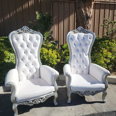 Pair of white and silver tufted throne chairs outdoors in sunlight, surrounded by shrubs and tree shade. (115 characters)