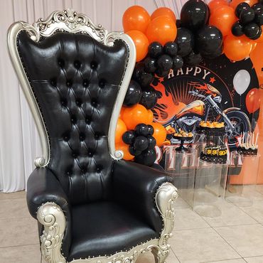 Black and silver tufted throne with ornate frame positioned next to an orange and black balloon arch at a motorcycle-themed birthday party for VIP seating.