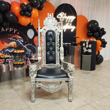 Tall silver throne chair with lion arms placed in front of a motorcycle-themed birthday backdrop with orange and black balloons for VIP guest seating.