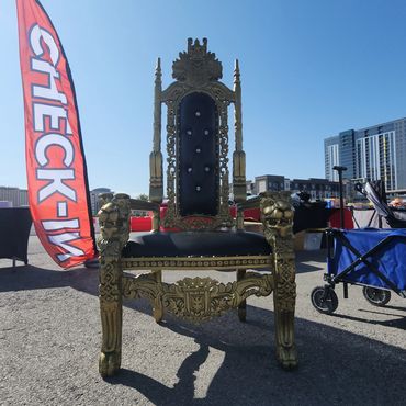Black tufted throne chair with detailed gold carving set up at an outdoor festival check-in area for branding or VIP guest photo opportunities.