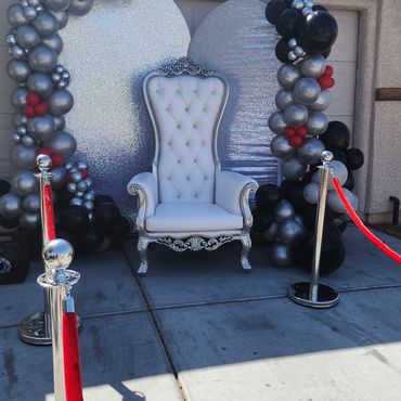 White and silver throne chair set outside with red carpet, stanchions, and black and silver balloon arch. (118 characters)