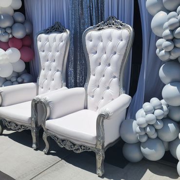 Two white and silver tufted thrones set against balloon columns and shimmering backdrop for outdoor event. (117 characters)
