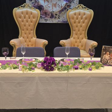 Pair of gold leather tufted thrones staged behind a floral-accented head table with purple décor for a formal wedding or anniversary celebration.
