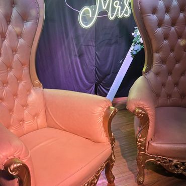 Tan gold tufted throne chairs with curved backrest placed under “Mr & Mrs” neon sign for romantic wedding sweetheart table setting.