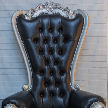 Black tufted high-back throne with silver carved frame photographed against a white brick backdrop for luxury event seating display.
