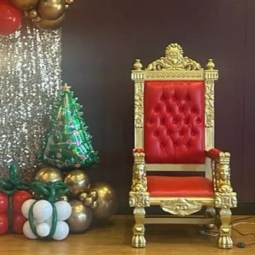 Gold and red throne flanked by balloon garland and decorated tree at indoor Christmas event or Santa setup.