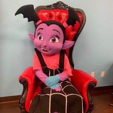 Cartoon vampire character sitting on red and black throne at an indoor event for kids or character meet-and-greet.