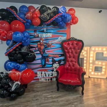Red tufted throne with black frame at a Spider-Man themed sweet 16 with balloon arch and marquee numbers.